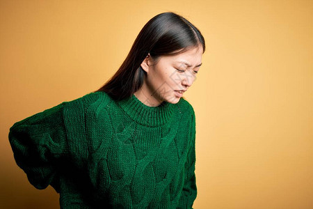 身穿绿色冬季毛衣黄色孤立背景的年轻美丽亚洲女背痛用手触摸背图片