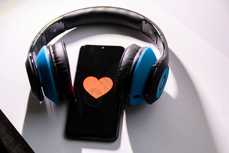 黑色屏幕上带有红心的黑色智能手机和无绳蓝色耳挂式耳机显示移动音乐流健康控制智能手机成瘾对音乐的图片
