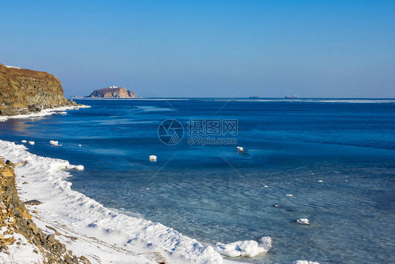 岩石海岸被白雪覆盖平静的水面明亮的蓝色远处是一座石岛晴朗阳图片