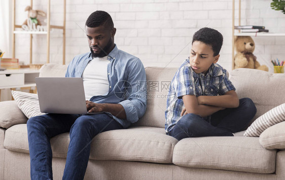 家长忽略概念小黑子被他父亲用笔记本电脑坐着暴躁和双手折叠在图片