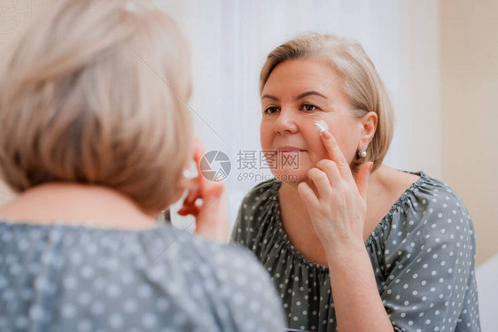 镜子前快乐健康的成熟女在脸上涂抹抗衰老保湿化妆霜图片