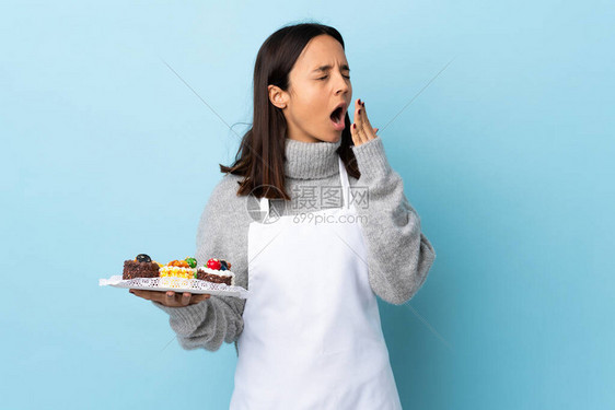 糕点厨师拿着一个大蛋糕在孤立的蓝色背景打哈欠用图片