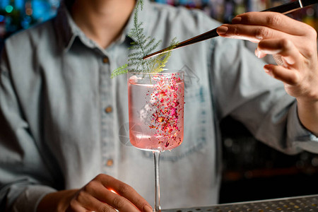 穿着玫瑰鸡尾酒装饰玻璃的年轻女男巴曼Barman使用tweezers用一小片绿色植图片