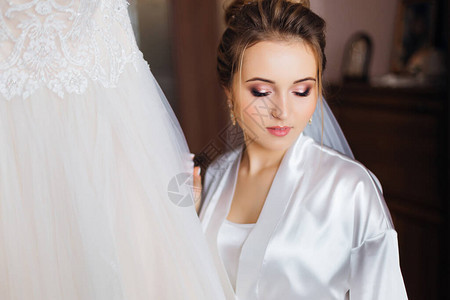 白浴袍上美丽的化妆品新娘在带蕾丝的婚纱旁边图片
