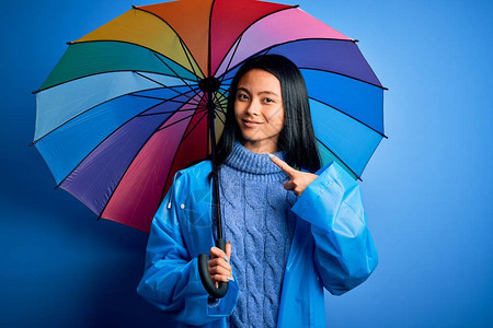 身穿雨衣在孤立的蓝色背景下撑伞的年轻女子非常高兴地用图片