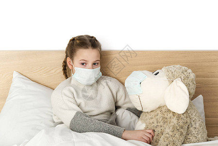 一个带着温度和的面具的生病悲伤的孩子躺在床上流感冒细菌在卧室里带玩具的女孩戴上防护面具图片