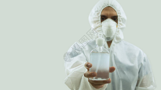 戴面罩和白色生物防护服的医生手握着一瓶白底浅地表面的凝胶酒精图片