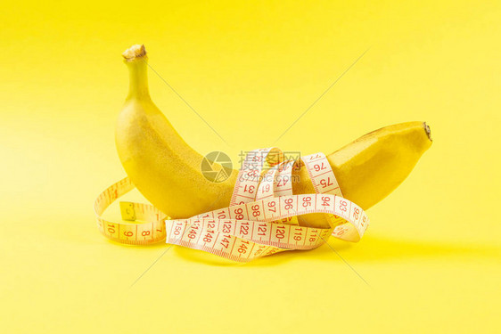 用胶带测量数字的香蕉厘米尺在水果周围旋转胶带缠绕在黄色背景上孤立的香蕉上减肥健康食品和苗图片