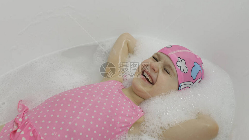 迷人的四岁女孩穿着粉红色泳装和泳帽用沐浴泡沫洗澡躺在浴缸里可爱的金发孩子的卫生可爱的女孩微笑浴室里图片