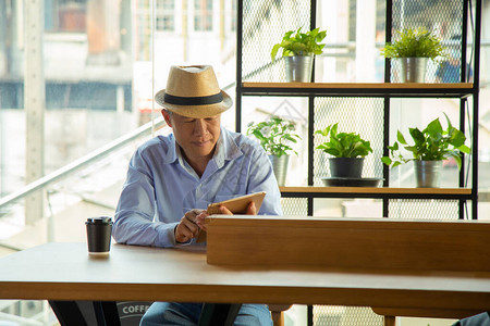中等特写肖像中年亚洲男子在休闲浅蓝色shite和牛仔裤子和凉爽的帽子坐在一边咖啡桌酒吧使用平板电脑上的应用程序与植物架与城市背景图片