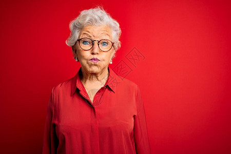 身穿休闲衬衫和眼镜的高级美丽灰发女人在红色背景上鼓着脸颊图片