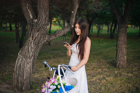 身穿白裙子的女孩和手拿着智能手机的背心站在一辆自行车旁边图片