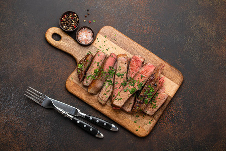 烤或油炸的优质大理石肉牛排Ribeye切片并在木制砧板上与餐具一起食用在质朴的棕色混凝土背景上切成片的多汁熟牛图片