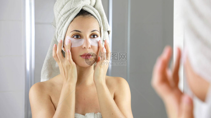美丽的女人应用抗疲劳眼罩在浴室的镜子里看着自己皮肤护理女孩在眼睛下方触摸织物面膜贴图片