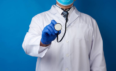 穿白色大衣的医生蓝色乳胶手套和面罩将听诊器放在蓝色背景上图片