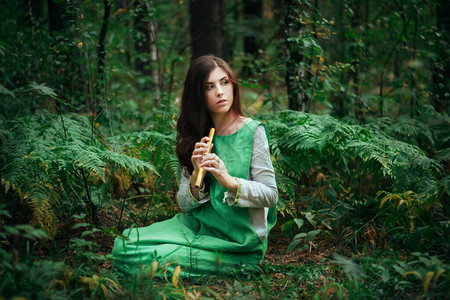 一位身穿绿色连衣裙的中世纪妇女坐在蕨类灌木丛中吹奏着木笛一个女孩在阴暗的森林里用手工烟斗演奏音乐暮色奇幻森林图片