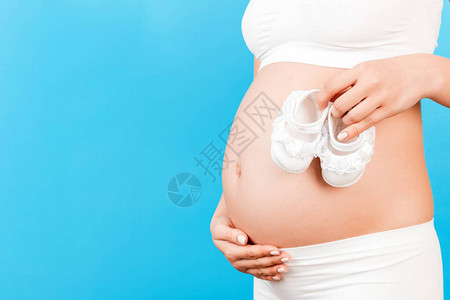 穿着白内衣的孕妇在蓝色背景下将女婴靴放在腹部对着肚子的刻画图象图片