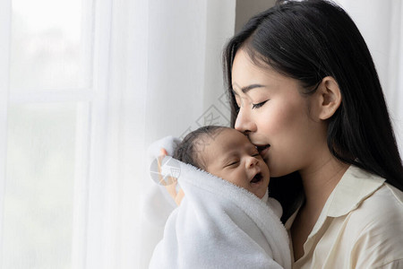 年轻的亚洲母亲在白床上房间亲吻她可爱的新生婴儿的肖像图片