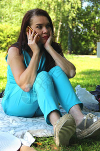 一个穿着绿松石裤套装的女人在夏日坐在绿色草坪上用手机说话图片