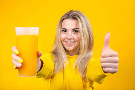 美丽的金发美女的肖像拿着橙汁杯举起拇指签好相片微笑的女孩在黄色图片