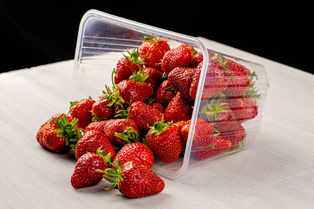 桌子上散着红熟多汁草莓的被推翻的透明塑料容器背景图片