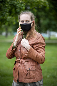 戴着黑色面具的美丽女人拿着电话站着穿着棕色夹克的女人公园里的女图片