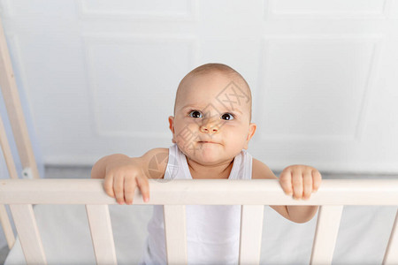 一个8个月大的微笑男婴身穿白色衣服站在儿童房的婴儿床里图片