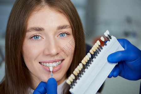 与牙科椅上微笑的女患者相匹配的牙色样本的近距离照片一位医生正在牙医诊所进行治疗前检查图片