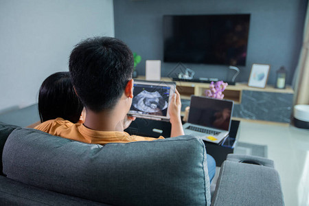 亚洲美丽的孕妇和丈夫在家中客厅沙发上看超声波扫描图片