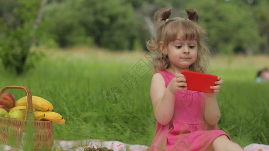 周末野餐草地上的女孩带着装满水果的篮子在手机上玩网络游戏儿童孩子使用智能手机在互联网上看电影聊天图片
