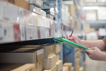 亚洲经理人使用剪贴板文件和笔在仓库货架上的纸板箱中对产品管理进行盘点背景图片