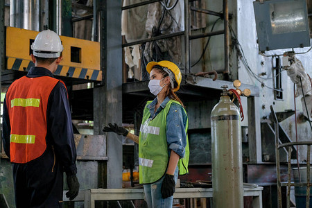 戴着卫生面具的工厂人男女在工作区的大机器前一起讨论工业企良好管理体系的概念图片
