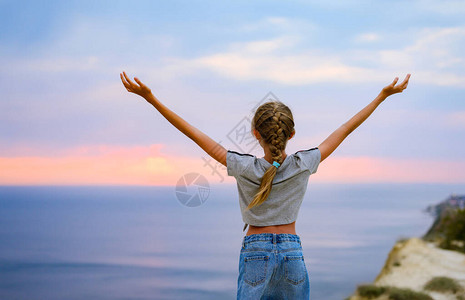女孩高举双手站在山边快乐的小女孩看着日落和大海图片