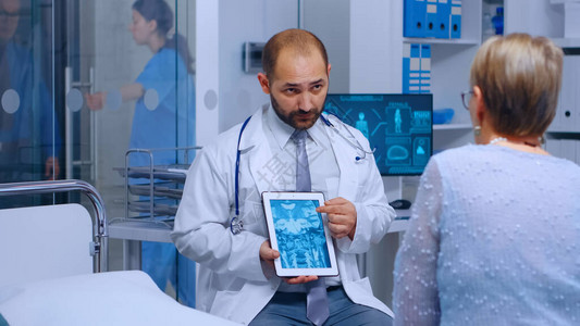 执业医生向坐在现代私人诊所病床上的高级退休妇女展示数字平板电脑上的放射学扫描医学显示屏幕图片