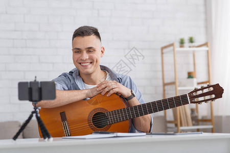 在线音乐课带着吉他微笑的家伙在室内客厅自由空间看智能手机相背景图片