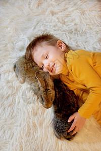 一个小男孩躺在温暖的地毯上与两只灰兔一起躺着时图片