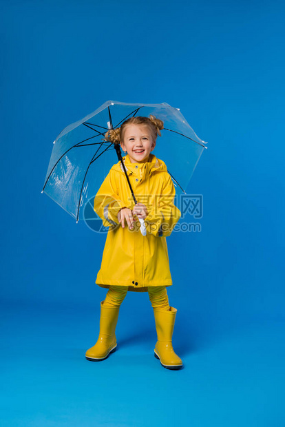 穿着雨衣和橡皮靴的快乐女孩站在蓝色背景上图片