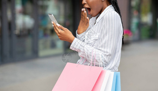 震惊的非裔美国妇女张嘴看智能手机持有纸包户外全景自由空间等图片