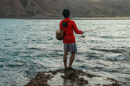 渔夫在低潮下钓鱼从水里有鱼棒的家伙的背面看到紧贴着身穿红色连帽衫背上挂着老鼠篮子的图片