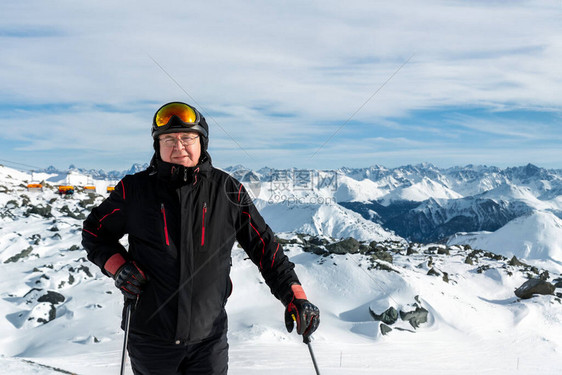 身着滑雪头盔护目镜和黑色西装的活跃资深老者站在山峰上享受冬季极限运动活令人惊叹的全景山图片