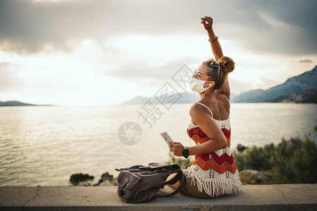 一名戴着防护面具的快乐年轻女子在海边探索日冕大流行期间用智能手机图片