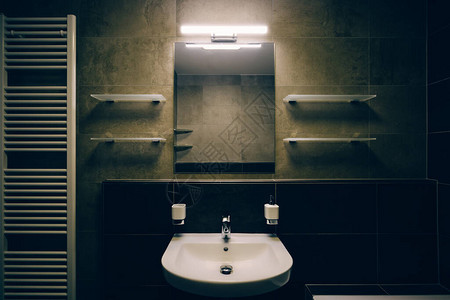 浴室墙有白色洗脸盆和镀铬水龙头墙上有镜子玻璃架子皂液器陶瓷杯和明亮的发光灯在左侧可以看到加图片