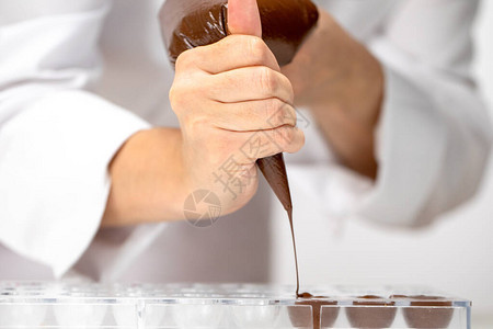 特写巧克力师将黑巧克力倒入糖果模具中厨师在白色围裙使用糕点袋填充塑料模具与热融化的巧克力制作自制巧克力糖背景图片
