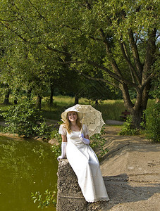 穿着白色历史服装的年轻女士戴着白色雨伞的白帽子坐在湖边公园里图片