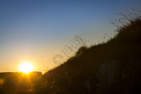 日落时在山上的岩石后面有草剪影的太阳图片