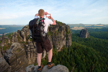 背着包的游客用双手的指制作框架背着大背包的徒步旅行者站在森林上方的岩石观景点阳光明媚的春天破图片