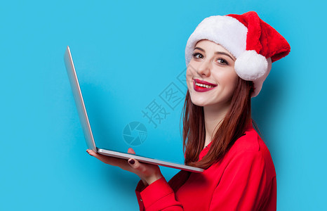 一名穿着圣诞老人帽子和蓝背景笔记本电脑的年轻红图片
