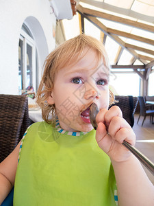 两岁金发的金发婴儿的脸嘴里塞着绿边饼和银金属勺子在餐厅里背景图片