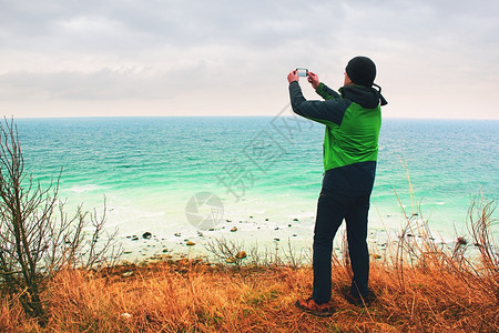 穿绿色防水夹克的人用智能手机拍摄了海岸风景的照片海面礁图片