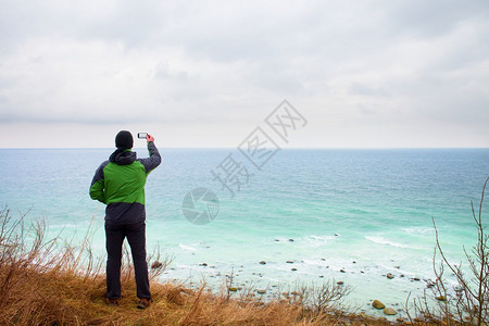 穿绿色防水夹克的人用智能手机拍摄了海岸风景的照片海面礁图片